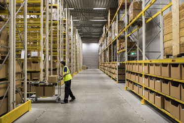 Full length side view of young warehouse worker pushing cart on aisle in industrial building - MASF09167