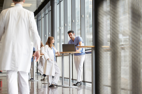 Mittelteil eines gehenden Arztes, während Mitarbeiter des Gesundheitswesens über einen Laptop auf dem Flur eines Krankenhauses diskutieren, lizenzfreies Stockfoto