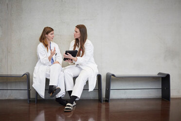 Selbstbewusste Ärztinnen in voller Länge, die über ein digitales Tablet diskutieren, während sie im Krankenhaus an der Wand sitzen - MASF09205