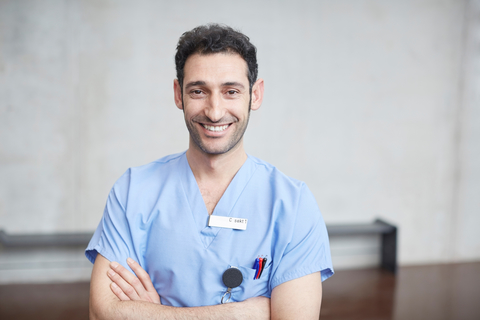 Porträt eines lächelnden jungen Krankenpflegers in blauem Kittel, der mit verschränkten Armen an der Wand eines Krankenhauses steht, lizenzfreies Stockfoto