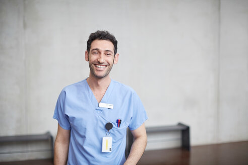 Portrait of smiling young male nurse in blue scrubs standing against wall at hospital - MASF09219