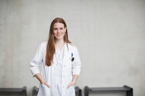 Portrait of smiling young female brunette doctor standing with hands in pockets at hospital - MASF09222