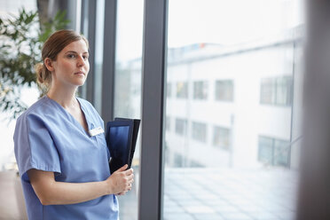 Thoughtful female nurse looking through window while standing in corridor at hospital - MASF09242