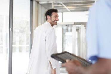 Lächelnder junger Arzt, der spazieren geht, während ein Krankenpfleger in der Lobby eines Krankenhauses ein digitales Tablet hält - MASF09271
