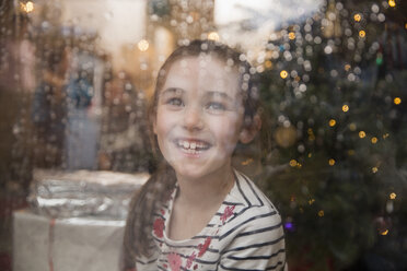 Porträt glückliches Mädchen am nassen Fenster im Weihnachtswohnzimmer - HOXF03799