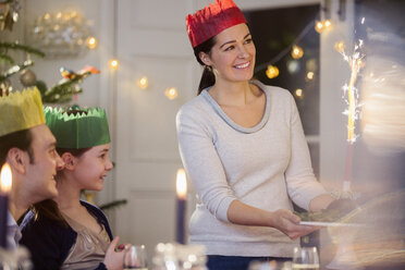 Glückliche Mutter in Papierkrone serviert Weihnachtspudding mit Feuerwerk bei Kerzenlicht am Tisch - HOXF03833