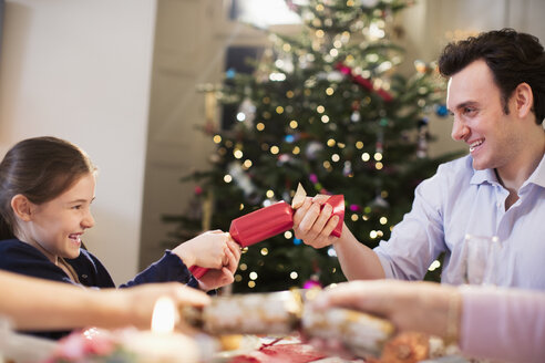 Vater und Tochter ziehen am Weihnachtstisch an einem Cracker - HOXF03889