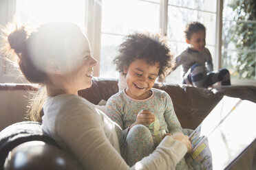 Happy mother and daughter cuddling, reading book on sofa in sunny living room - HOXF03917