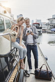 Older man with camera and young woman standing with travelling bags on jetty next to yacht - RORF01552