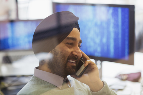 Indischer Computerprogrammierer mit Turban, der im Büro mit einem Smartphone telefoniert, lizenzfreies Stockfoto