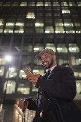 Lächelnder Geschäftsmann mit Smartphone, der unter einem Hochhaus bei Nacht steht - CAIF21951