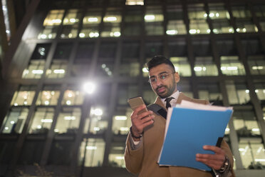 Geschäftsmann mit Papierkram und Smartphone unter städtischem Hochhaus bei Nacht - CAIF21972