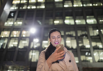 Lächelnde Geschäftsfrau mit Smartphone unter städtischem Hochhaus bei Nacht - CAIF21977