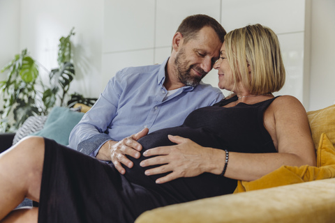 Reifer Mann und seine schwangere reife Frau sitzen auf der Couch und berühren ihren Bauch, lizenzfreies Stockfoto