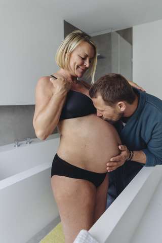 Reifer Ehemann küsst den Bauch der werdenden Mutter im Badezimmer, lizenzfreies Stockfoto