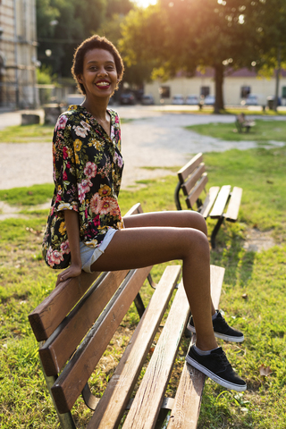 Porträt einer lächelnden jungen Frau, die auf einer Bank in einem Park sitzt, lizenzfreies Stockfoto