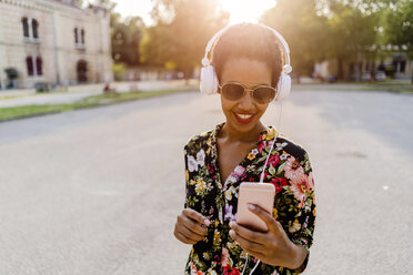 Glückliche, modische junge Frau mit Kopfhörern und Smartphone im Freien bei Sonnenuntergang - GIOF04319
