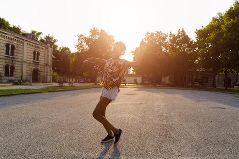 Glückliche modische junge Frau mit Kopfhörern tanzt im Freien bei Sonnenuntergang - GIOF04322