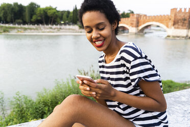 Lächelnde junge Frau sitzt auf der Mauer am Flussufer und benutzt ein Handy - GIOF04327
