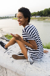 Smiling young woman sitting on wall at the riverside using cell phone - GIOF04328