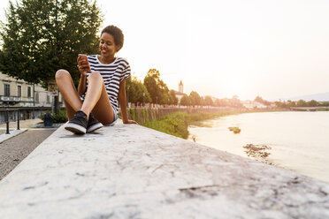 Smiling young woman sitting on wall at the riverside using cell phone - GIOF04329