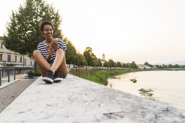 Smiling young woman sitting on wall at the riverside using cell phone - GIOF04330