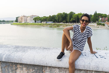 Porträt einer lächelnden jungen Frau mit Sonnenbrille, die am Flussufer sitzt - GIOF04332