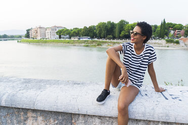 Lächelnde junge Frau mit Sonnenbrille am Flussufer sitzend - GIOF04333
