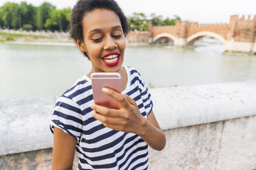 Smiling young woman using cell phone at the riverside - GIOF04337