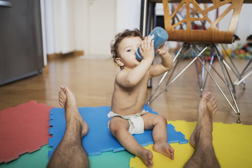 Ein kleiner Junge in einer Windel sitzt mit seinem Vater zu Hause auf dem Boden und trinkt Wasser - AZOF00008