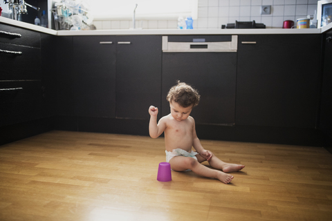 Baby boy sitting on floor in the kitchen playing with plastic cup stock photo
