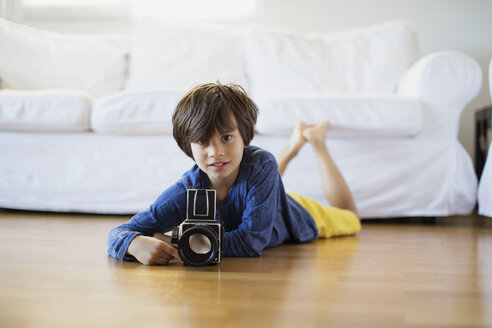 Porträt eines Jungen, der zu Hause mit einer altmodischen Filmkamera auf dem Boden liegt - AZOF00017