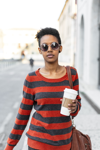 Porträt einer jungen Frau mit Kaffee zum Mitnehmen, die eine Sonnenbrille und einen gestreiften Pullover trägt, lizenzfreies Stockfoto