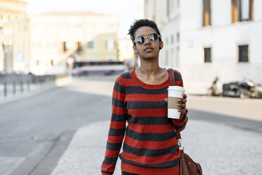 Porträt einer jungen Frau mit Kaffee zum Mitnehmen, die eine Sonnenbrille und einen gestreiften Pullover trägt - GIOF04343