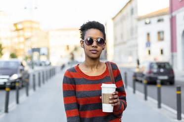 Porträt einer jungen Frau mit Kaffee zum Mitnehmen, die eine Sonnenbrille und einen gestreiften Pullover trägt - GIOF04344