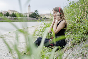 Lächelndes jugendliches Mädchen mit Kopfhörern, das am Flussufer Musik hört - GIOF04374