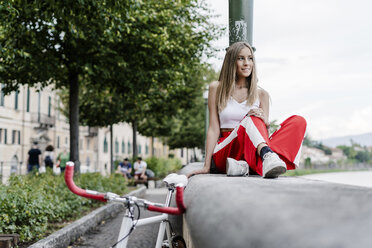 Lächelndes Teenager-Mädchen sitzt auf einer Mauer am Flussufer neben einem Fahrrad - GIOF04385