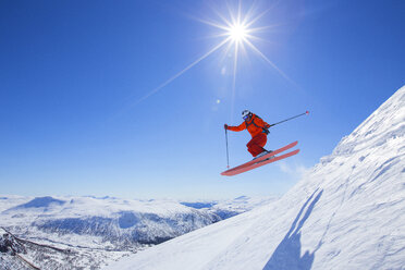 Ein männlicher Freerider in einem roten Anzug springt von einem Schneekamm. Die Sonne scheint, der Himmel ist blau. - AURF04032