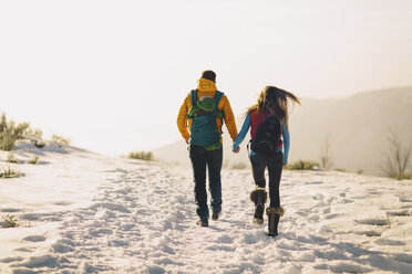 Ein junges Paar hält sich an den Händen und wandert vor einer winterlichen Bergkulisse und einem hellen Sonnenuntergang. - AURF04165