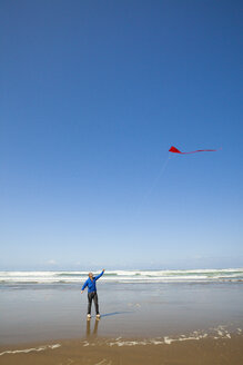 Ein junger Mann schaut bei einem Strandbesuch an der Küste von Oregon zu seinem roten Drachen am Himmel hinauf - AURF04211