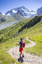 A colorful dressed male hiker is descending curved mountain trail in a green summer landscape with glaciers in the background. - AURF04397