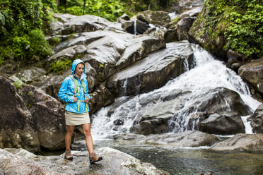 Ein Porträt einer Frau, die vor einem Wasserfall und einem Bach in einem Wald in Puerto Rico steht und eine bunte Regenjacke trägt. - AURF04549