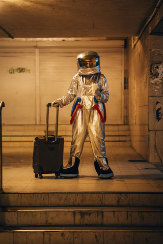 Raumfahrer in der nächtlichen Stadt, der mit Rollkoffer und Kaffee zum Mitnehmen in einer Unterführung steht, lizenzfreies Stockfoto
