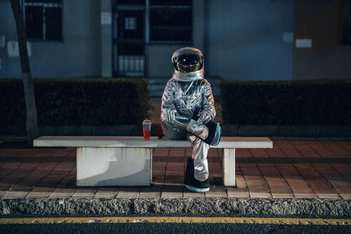 Spaceman sitting on bench at a bus stop at night with soft drink - VPIF00733