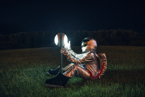 Raumfahrer sitzt nachts auf einer Wiese und staunt über eine Lampe, lizenzfreies Stockfoto