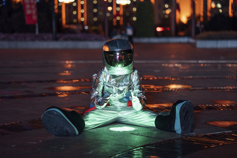 Raumfahrer, der nachts an einer Lampe auf einem Stadtplatz sitzt und sich die Hände wärmt, lizenzfreies Stockfoto