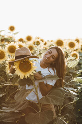 Portrait of a young laughing woman with straw hat, standing in a field of sunflowers - OCAF00362