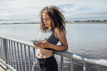 Junge sportliche Frau mit Kopfhörern und Smartphone am Flussufer - VPIF00782