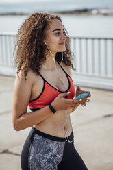 Smiling young athletic woman with smartphone and earbuds at the riverside - VPIF00785