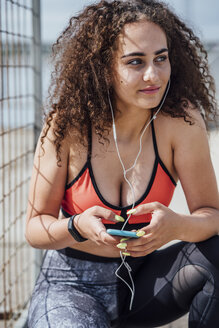 Lächelnde junge sportliche Frau mit Smartphone und Kopfhörern, die im Freien hockt - VPIF00789